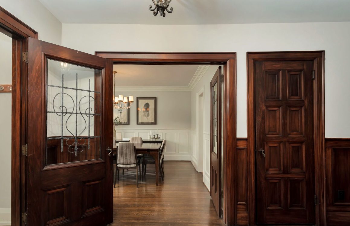 Walnut-panelled foyer with a glazed door and wrought-iron grille, opening onto a dining room with white wainscoting and a candle chandelier